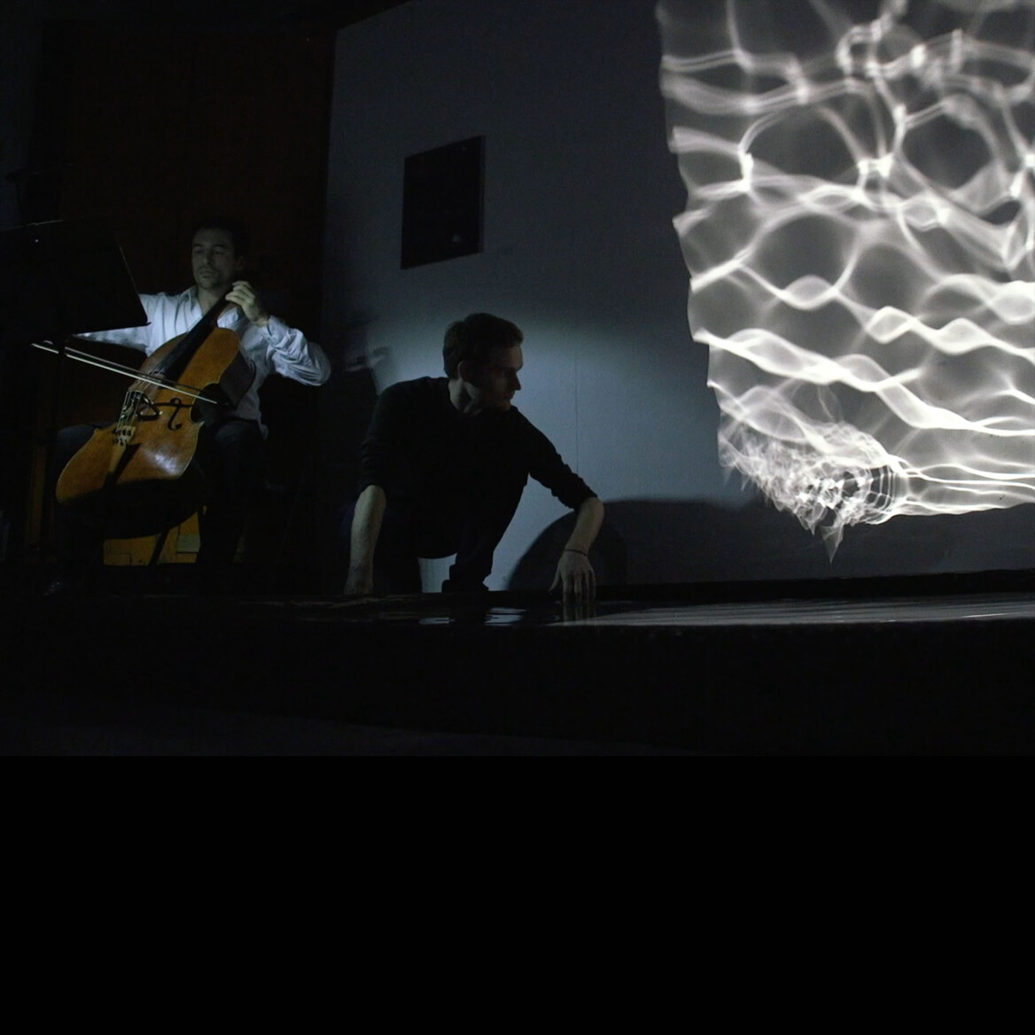 Man playing a cello to the left, another man crouching down with his hand in large square pool of water, with reflected patterns of the pool projected onto the top right of the image, showing beautiful patterns of light off the water.