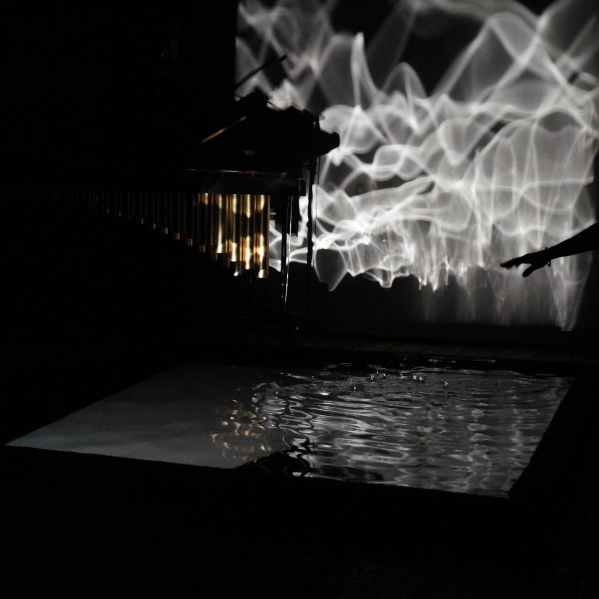 Kate interacts with a shallow square pool of water as sound vibrations create ripples in the water which are reflected onto a wall