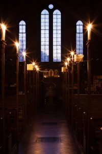 Long-exposure photo of Kate lighting candles, where a visible light trail left by the match can be seen as the sun sets outside the stained glass windows