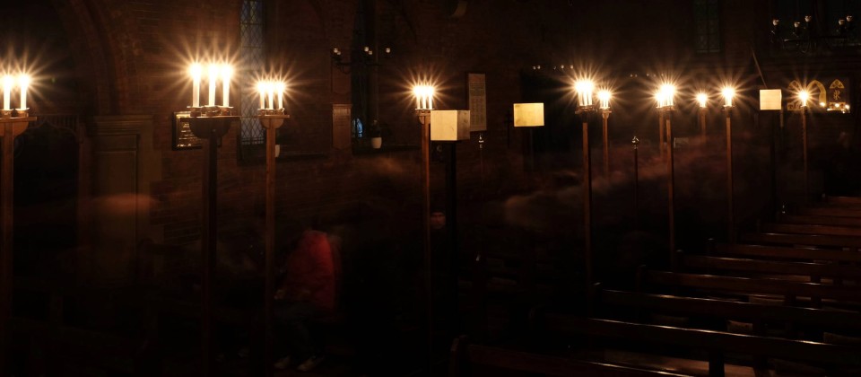 Photo showing the dark interior of the church, candles along the central aisle burning bright as the crowd pass through the installation in slow exposure