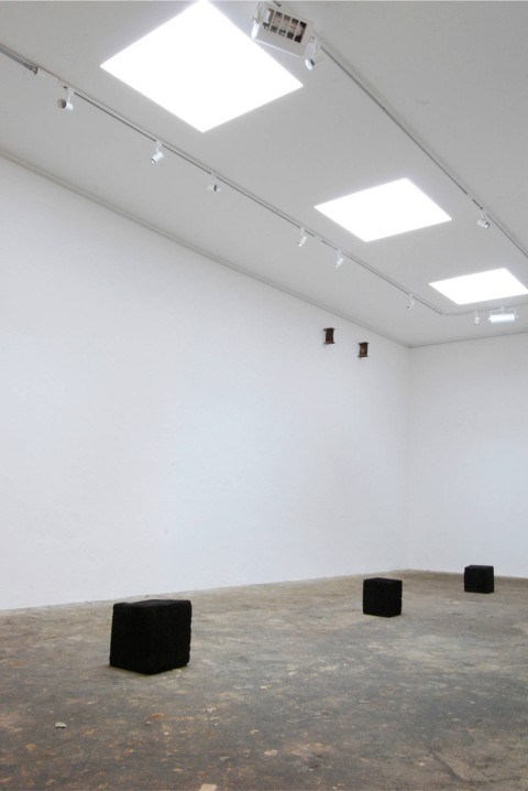 Photo documenting the interior of the exhibition space, which shows three large cubes of burnt oak arranged on the gallery floor in a line under three cubic skylights.