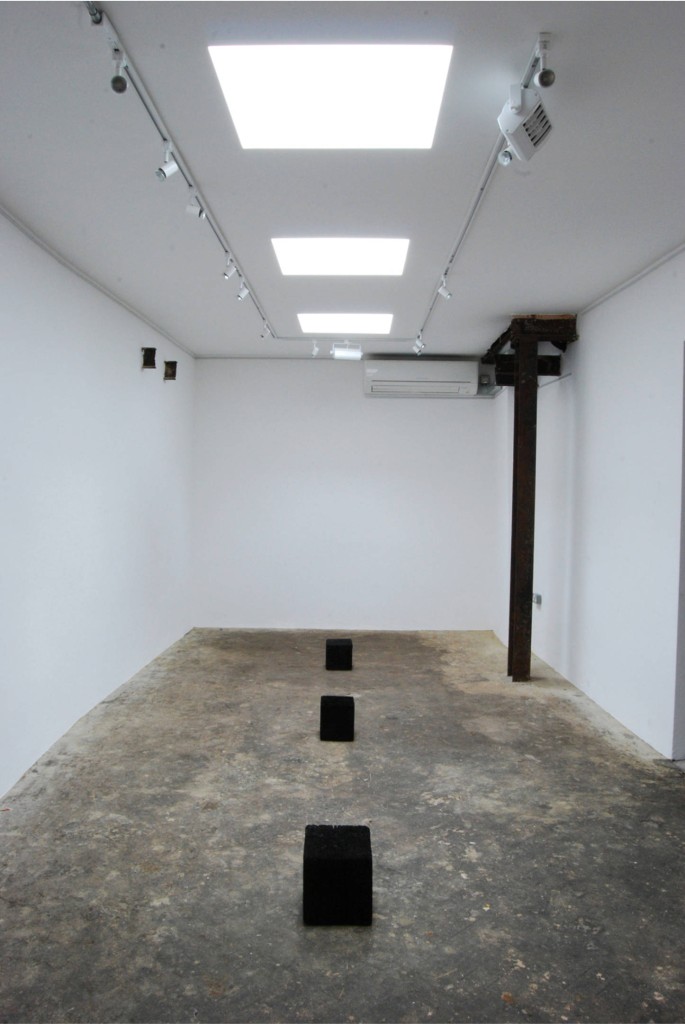 Photo documenting the interior of the exhibition space, which shows three large cubes of burnt oak arranged on the gallery floor in a line from a straight-on perspective