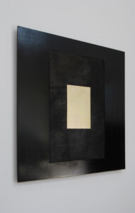 Side view of the artwork "Trinity", which has a small oblong gold shape in the middle, surrounded by a burnt black wood rectangle, which is surrounded by a black lacquer larger rectangle.
