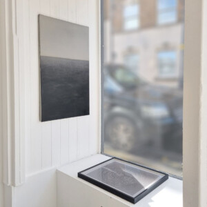 A canvas depicting a dark sea and blank sky on the wall and a framed photogram lying on the window sill