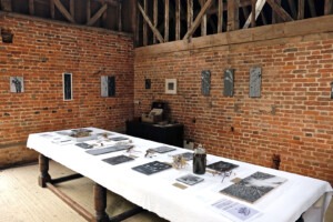 Exhibition shot of long table with many works on it along with the flora the works were made from. The brick walls also have works hanging