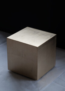 A medium sized crisp edged cube covered in immaculately flat pale gold leaf. The three faces seen have varying amounts of light on them. The cube is sitting, floating just above a white tissue floor and fades into a black area behind it.