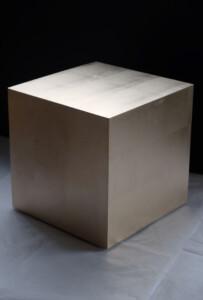 A large, crisp edged cube covered in immaculately flat pale gold leaf. The three faces seen have varying amounts of light on them. The cube is sitting, floating just above a white tissue floor and fades into a black area behind it.