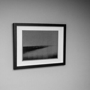 black framed grey and black photograph with sea and sea wall fading into distance, in stages